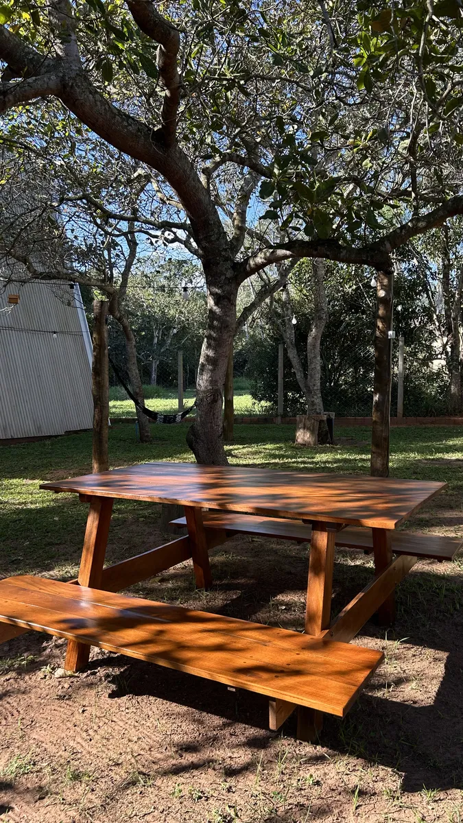 Mesa de piquenique de madeira à sombra das árvores, com redário ao fundo